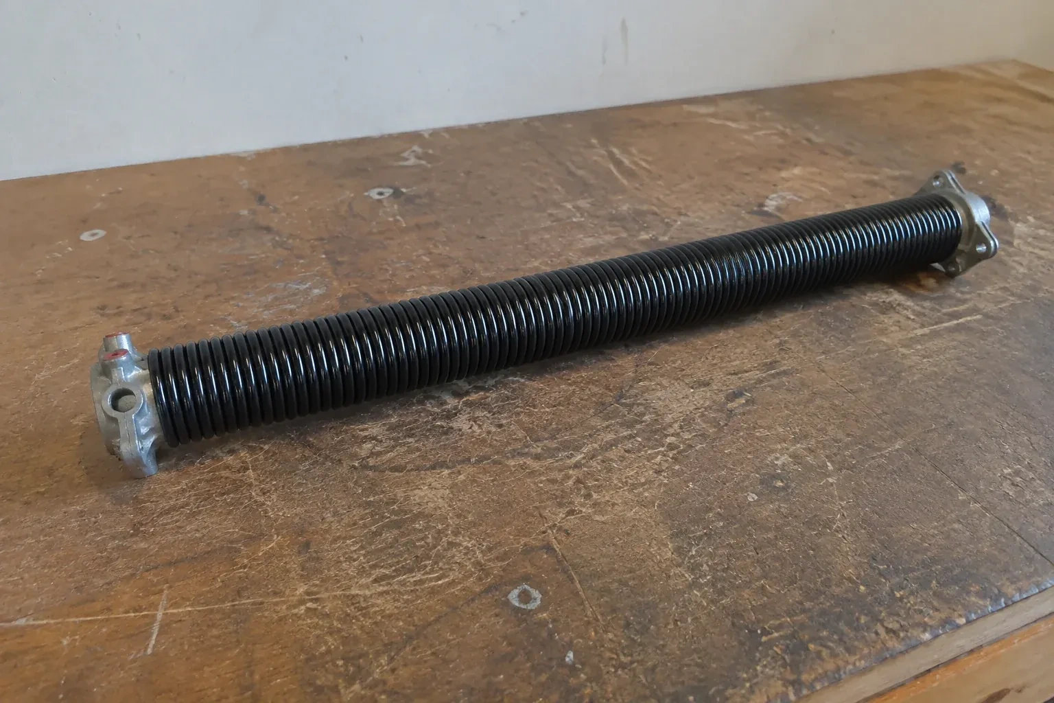 Detached black coil spring with metal end fittings on a workbench. — Torsion Spring Replacement, Toronto