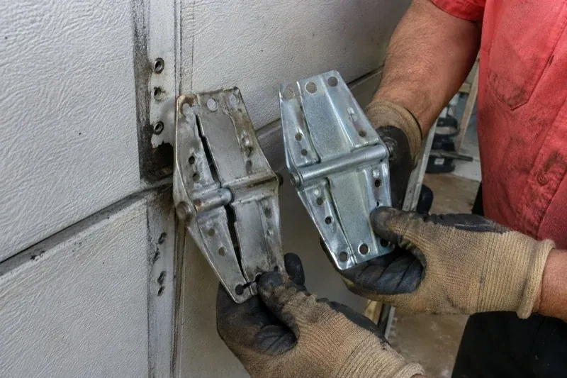Gloved hands holding two metal garage-door hinges side by side, one discolored with a visible crack and the other bright silver. — Garage Door Hinge Replacement, Toronto